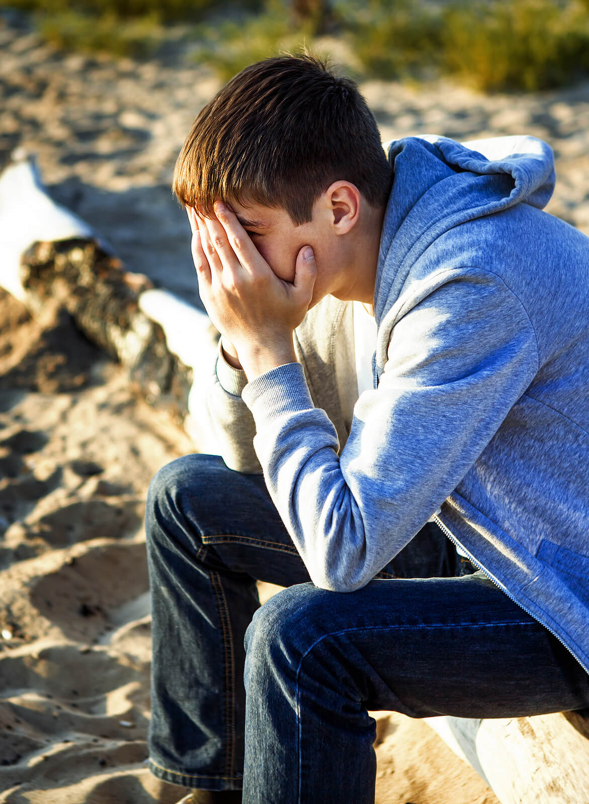 A teen boy sits down on a beach with his hands covering his face. Wondering if Therapy for Teens in Arlington, MA can help your teen? Speak with a teen therapist in MA to see how trauma in teens is affecting your teen.
