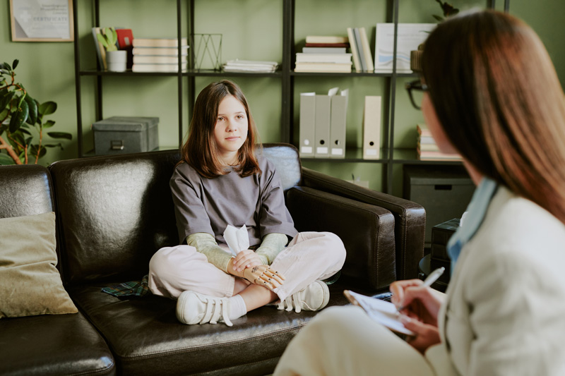 A teen girl client sits down with her female therapist on a couch. Want to learn how Therapy for Teens in Arlington, MA can help your teen? Speak with a teen therapist to learn how therapy helps trauma in teens in Arlington, MA.