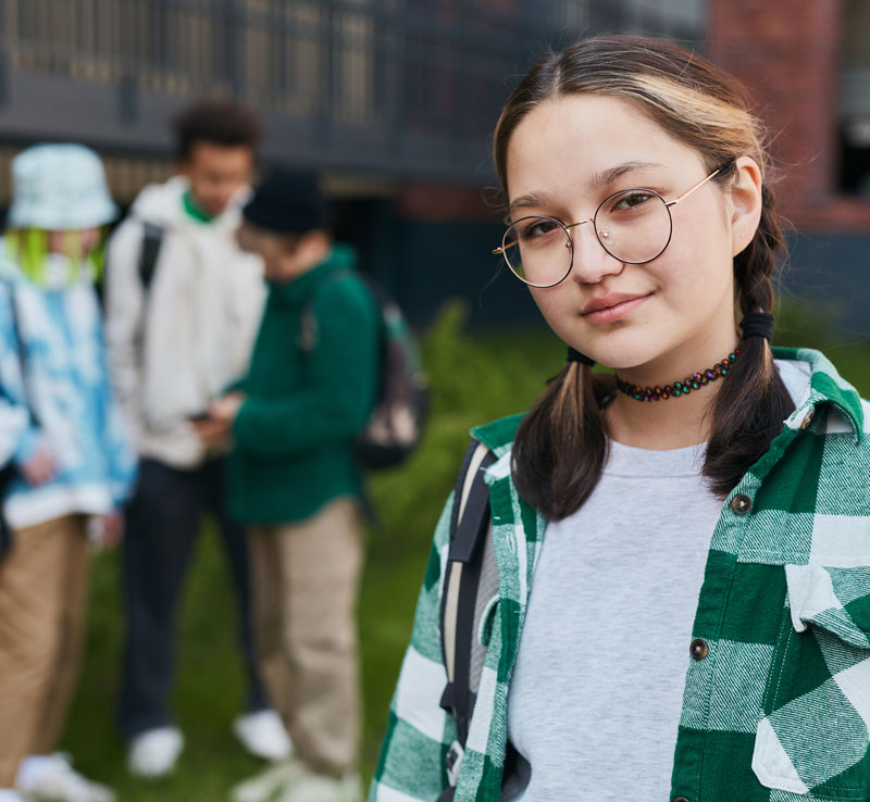 young teenage girl looking at the camera with a half smile while other teenagers are in the background talking. Teens find mental health support during Therapy for Teens at BainHWC