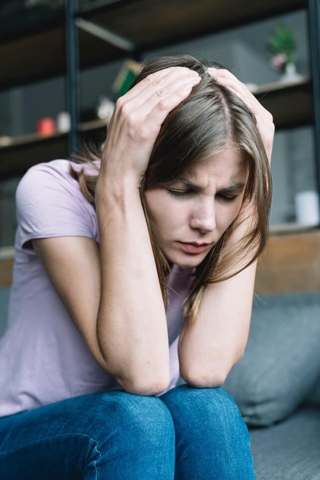 A teen girl sits on a couch holding her head with her hands. Learn more about how Therapy for Teens in MA can help your teens with OCD in Arlington, MA navigate their symptoms.