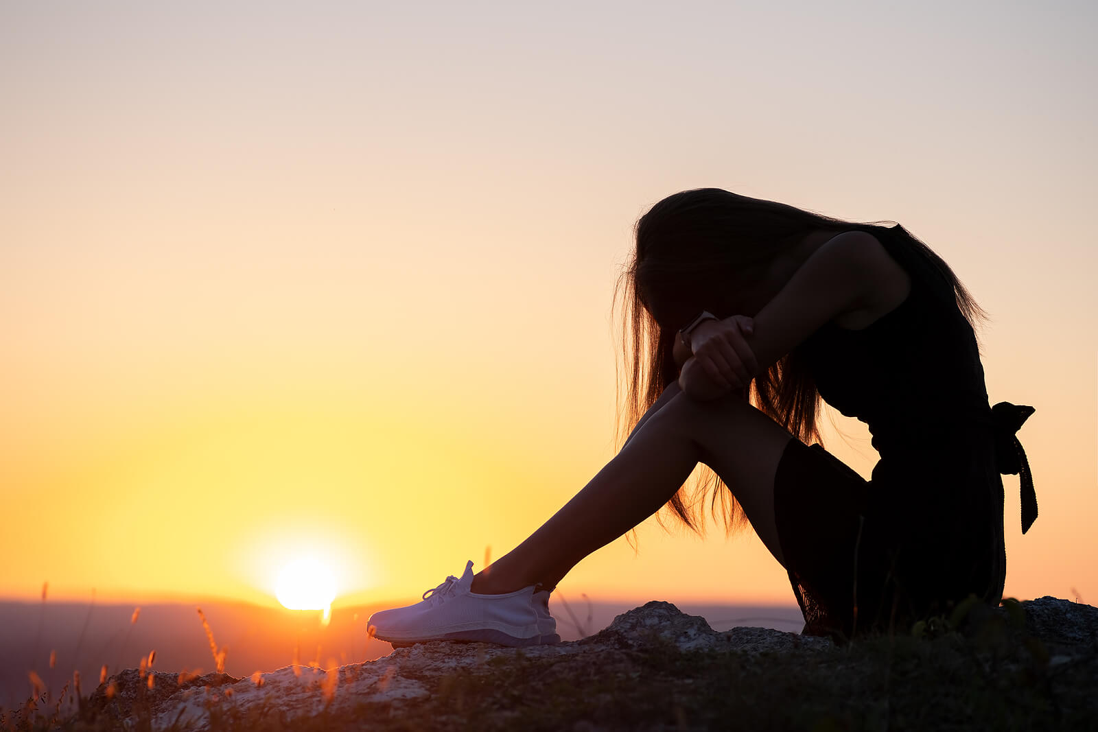 a silhouetted teen girl sits on a rock with the sunsetting. Wondering how to help your teens with depression in Arlington, MA? Speak with a teen therapist to see how Therapy for Teens in Arlington, MA can help. 