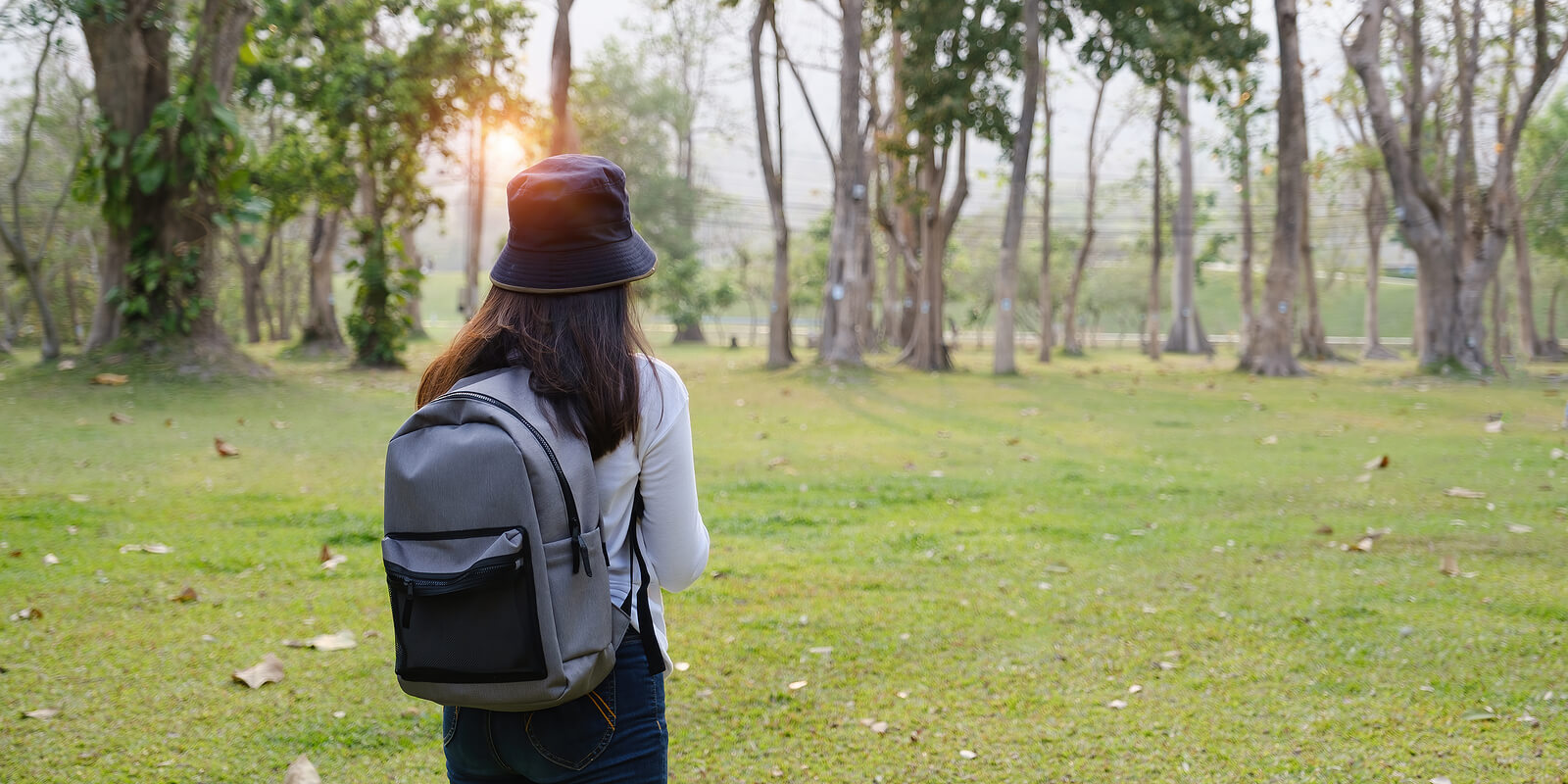 A teen with a bucket hat and a backpack on stands in a grassy area near trees. Want to learn how CBT works for teens with autism in Arlington, MA? Speak with a teen therapist in MA to see how therapy for teens can help them.