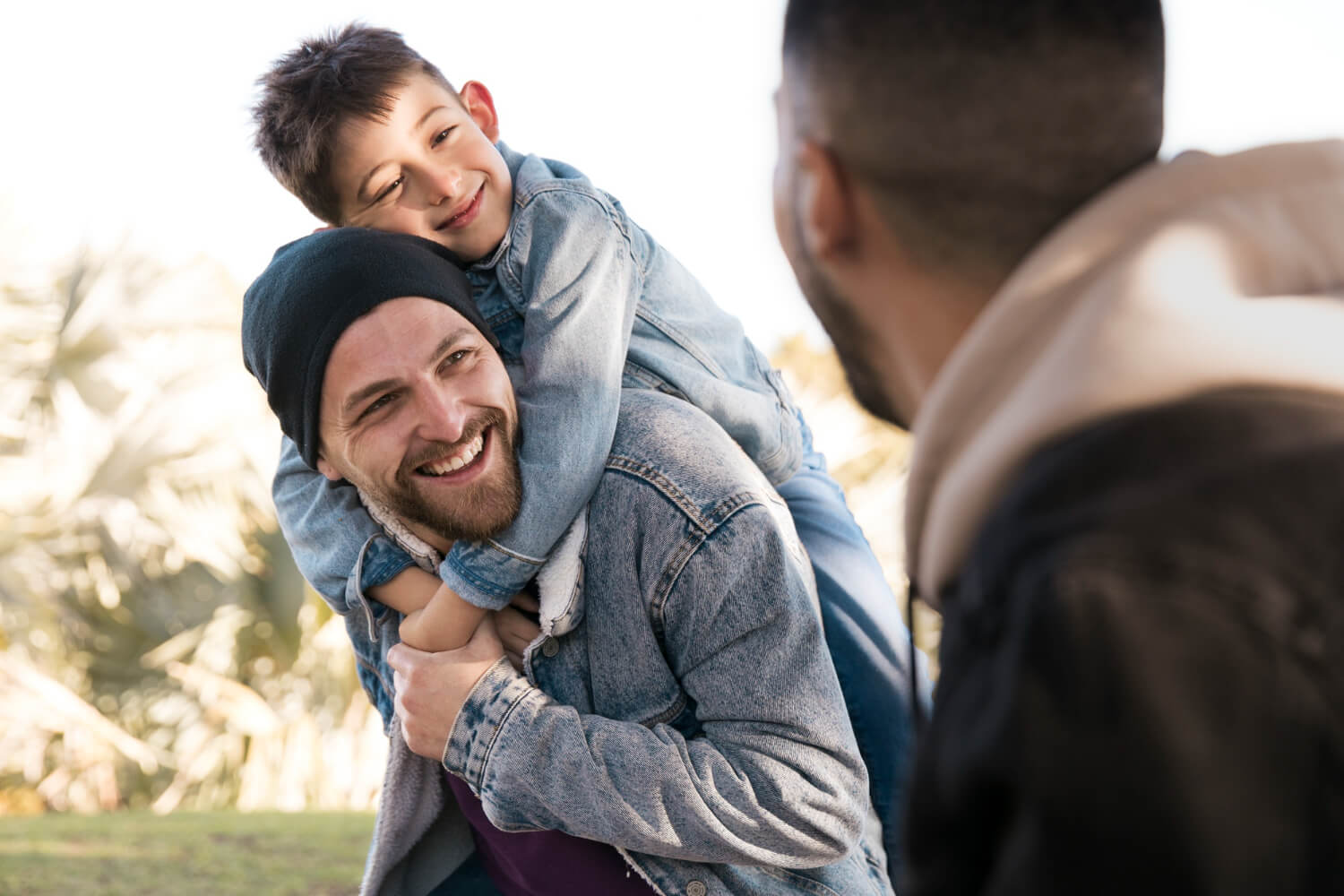 A man smiles while in the park with his husband and his son on his shoulders. Interested in learning how acceptance and commitment therapy for teens in Arlington, MA can help your teen navigate their emotions? Reach out to a teen therapist to learn more.