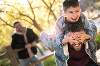 A man smiles while in the park with his husband and his son on his shoulders. Interested in learning how acceptance and commitment therapy for teens in Arlington, MA can help your teen navigate their emotions? Reach out to a teen therapist to learn more.