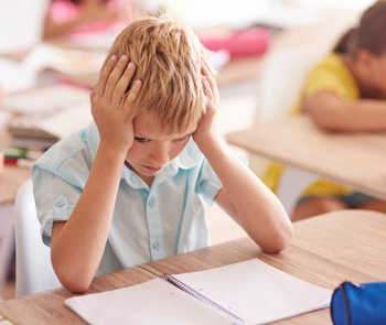 A teen boy holds his face with his hands while sitting at a desk in a classroom. Do you feel like your teen might benefit from Therapy for Teens in Arlington, MA? Speak with a teen therapist in MA to find out!