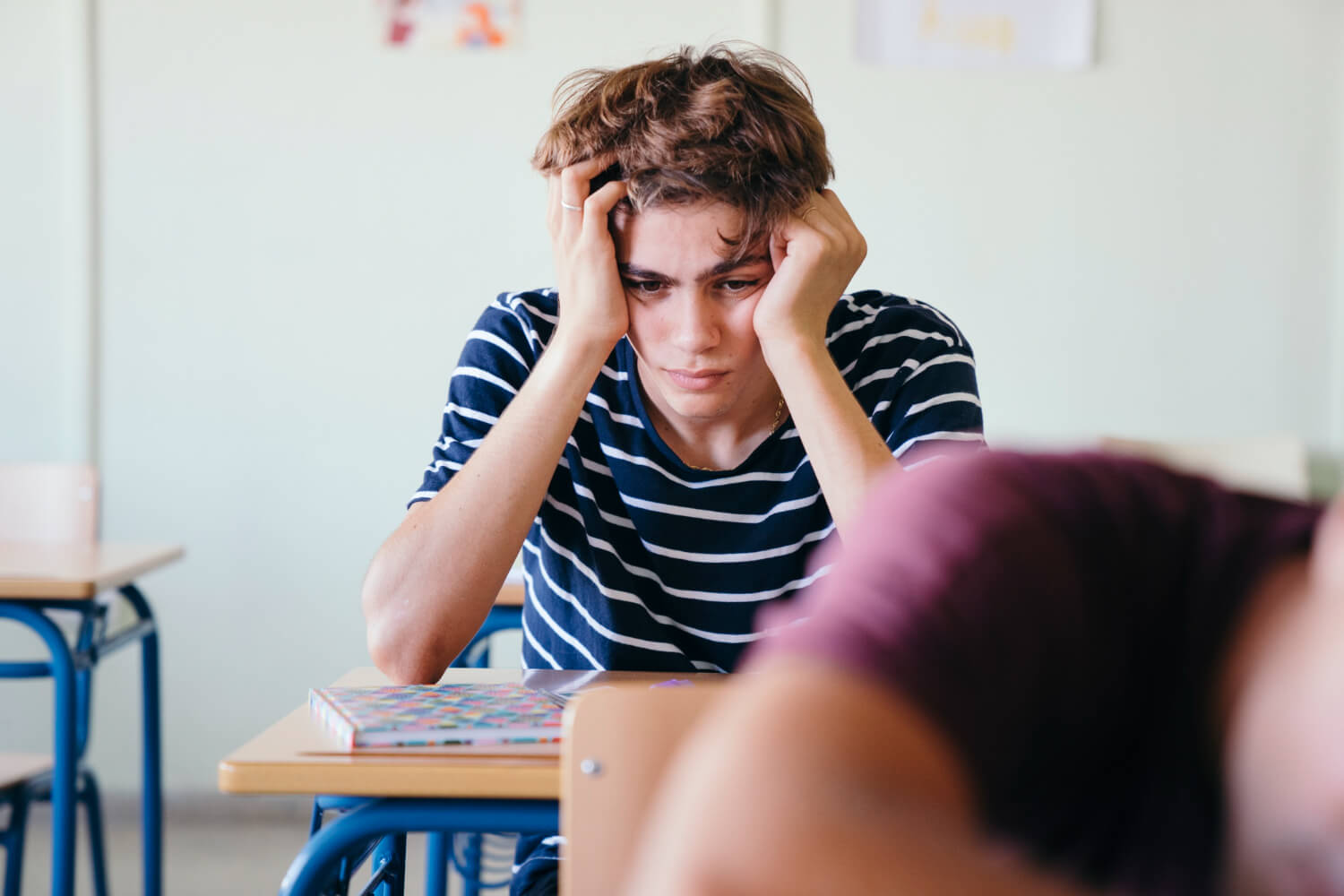 A teen boy holds his face with his hands while sitting at a desk in a classroom. Do you feel like your teen might benefit from Therapy for Teens in Arlington, MA? Speak with a teen therapist in MA to find out!