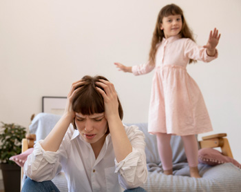 A mother touches her face with a stressed expression while her child is jumping on the bed. Search for ADHD treatment in Arlington, MA for parenting support and other services. They can offer therapy for parents Arlington, MA and more.