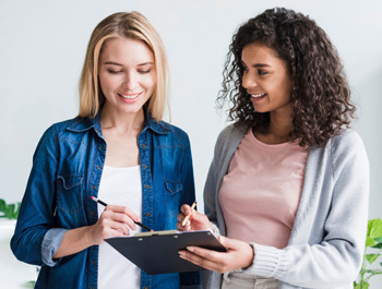 A woman with a pen and notepad stands with another woman as both converse. This could represent how parenting support in Arlington, MA can help parents and their children. Search for a teen therapist in Arlington, MA and search for ADHD treatment in Arlington, MA today.
