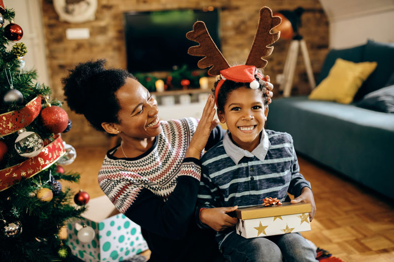 mother sits by the Christmas tree with her son while he tries on reindeer ears and smiles