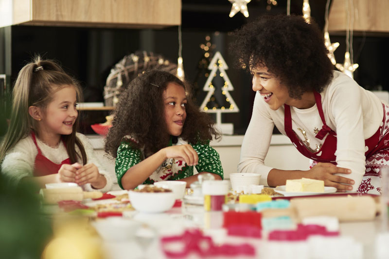 mother stands next to her daughters while they prepare Christmas cookies