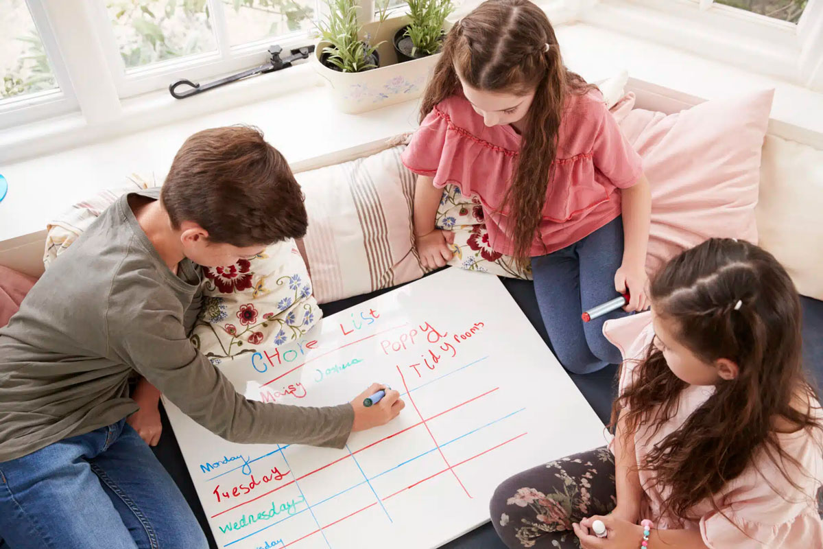 three children sit around a white board completing the chore list in multiple colors