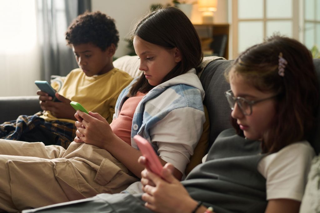 three young preteens sits side by side on a sofa each looking at their smart phones