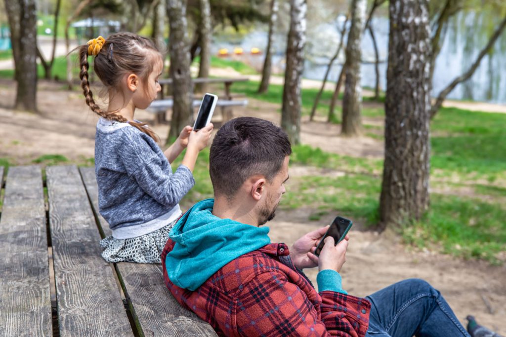 Father and daughter sitting together in a park near water, both using their mobile phones in silence