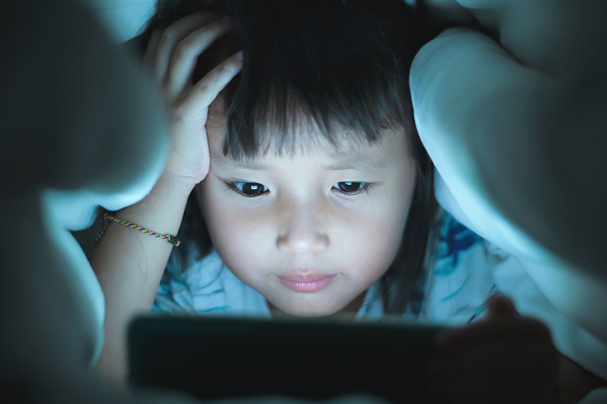 a young girl is sitting looking at a bright screen under the covers at night