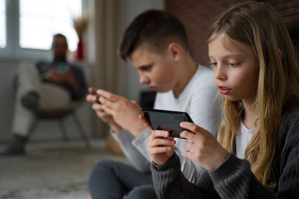 two preteens sit side by side looking at their smart phones